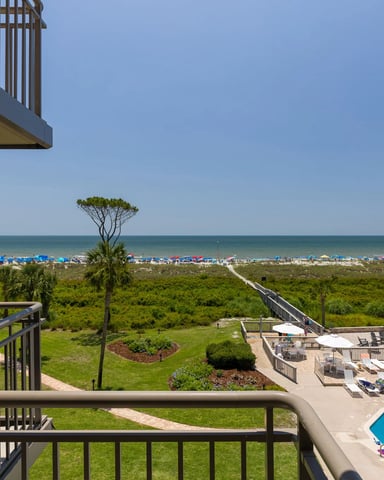 Oceanfront balcony view with pool, beach umbrellas, and palm trees overlooking the Atlantic coast