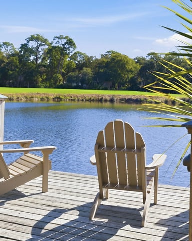 Wooden deck with Adirondack chairs overlooking a river, palm fronds in foreground, trees and green lawn beyond.