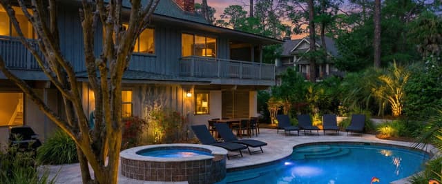 Modern home with illuminated pool and hot tub at dusk, surrounded by lounge chairs and palm trees